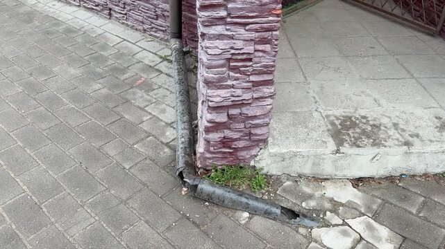 Cracked pipe by brick corner leaking onto pavement, corroded metal downspout detached from wall, small hole releasing water, moss