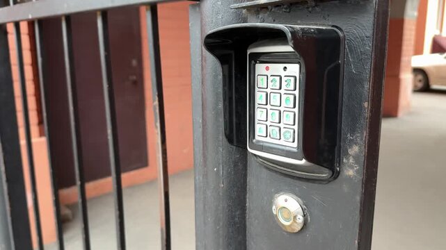 Analog intercom keypad on iron gate, weathered black frame, green backlit numeric buttons, round scanning key below keypad, brick