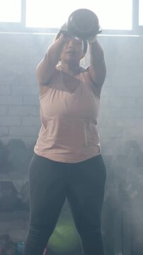 Vertical video: Gripping kettlebell, woman in pink top swinging for strength in gym with dumbbells