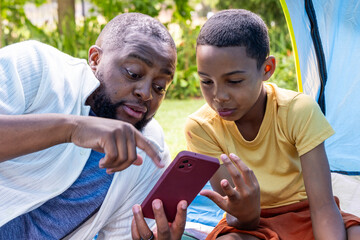 Fototapeta premium African American adult and teenage males leaning in, pointing at maroon smartphone at tent entrance