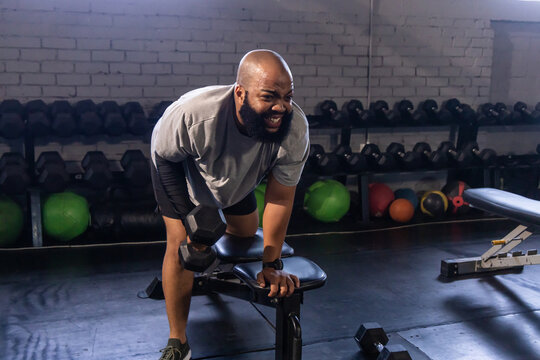 Adult African American man wearing gray tee pulling hex dumbbell in bent-over row on flat bench