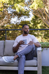 Obraz premium Mature African American man sitting on cushioned patio sofa holding ceramic mug, wearing T-shirt