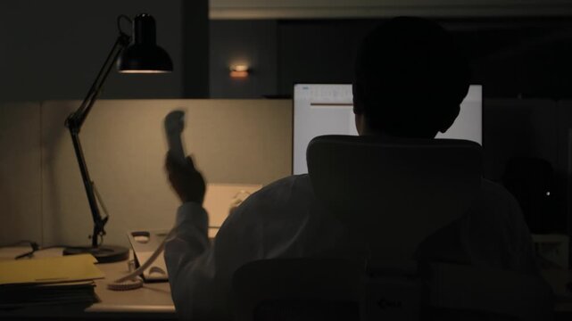A close-up of a stuffed teddy bear sitting on a desk beside a lamp in a dark office at night suggesting personal comfort during overtime work.