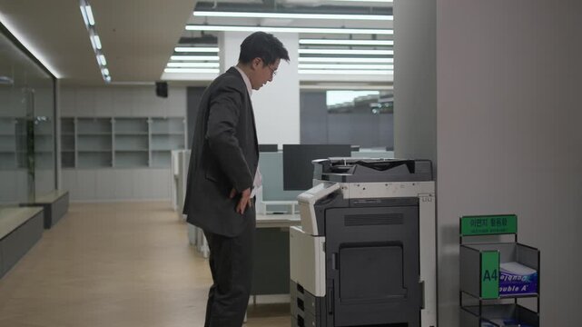 A businessman in a suit standing beside a printer machine in a modern office at night reviewing printed documents under bright fluorescent lighting.