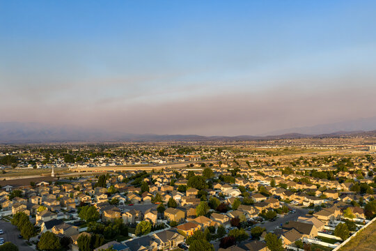 Aerial view of a suburban residential neighborhood with rows of houses, green trees, and local streets under a hazy sky caused by distant brushfire smoke, Lehi, Utah, United States.