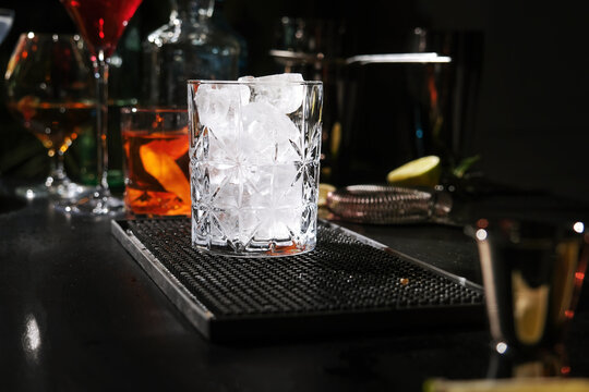 Crystal glass with ice cubes on bar mat against woman bartender background. Cooling drinks and bar setup concept.