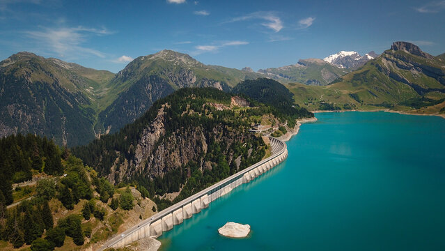 Aerial view of the Roselend Dam holding back the turquoise waters of the Roselend Reservoir, surrounded by lush green mountains and pine forests in Beaufort, Auvergne-Rhone-Alpes, France.
