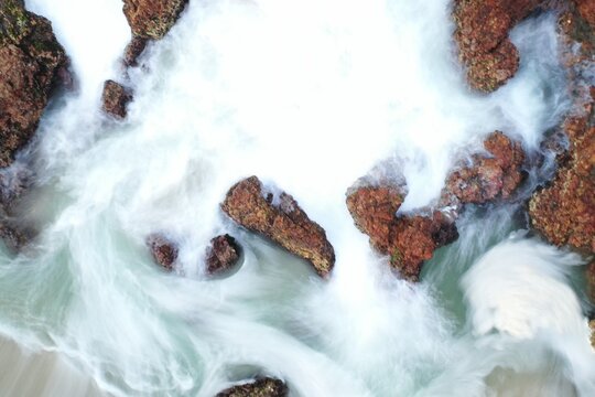 Aerial view of Flynns Beach with white sea foam swirling around rugged brown coastal rocks in Port Macquarie, New South Wales, Australia.