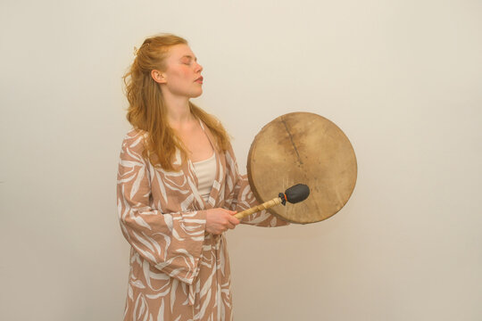Young woman plays a traditional frame drum during a sound healing session. Eyes closed in deep concentration, she expresses mindfulness, spirituality, rhythm and inner balance