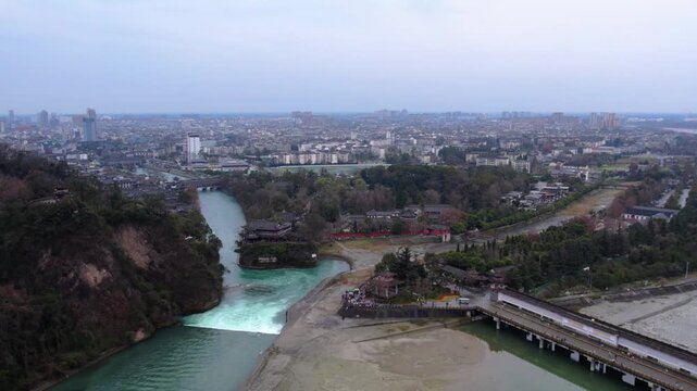 Aerial view of ancient water control works with river, bridge, trees and adjacent urban area under clear sky.