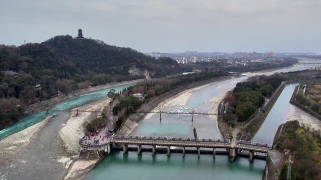 A landscape of a water diversion project with river channels, a bridge, forested hill, and distant urban area under overcast sky.