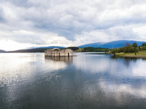 Aerial view of the submerged Church of St. John of Rila standing in the middle of a calm lake surrounded by rolling hills and a cloudy sky in Sliven, Bulgaria.
