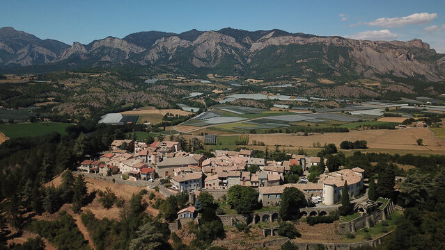 Aerial view of the historic village of Ventavon nestled among fields and greenhouses with a dramatic mountain range in the background in Ventavon, Provence-Alpes-Cote d'Azur, France.