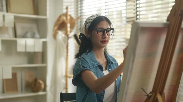 Young Asian woman artist sitting on stool painting blank canvas on wooden easel with palette and brush in stylish home art studio, full body creative lifestyle and hobby concept	