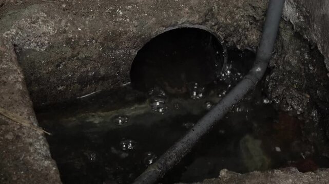 A close up shot of muddy stormwater rushing and bubbling into an open concrete drainage pit with a black pipe during a heavy downpour.
