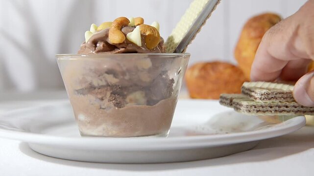 una copa de cristal con Helado de chocolate con chispas de chocolate blanc, almendras y nueces de la india encima, con panecitos y pared de madera en el fondo, una cuchara dentro de la nieve.