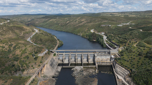 Aerial view of Fratel Dam spanning the Tejo River, surrounded by rolling green hills, winding roads, and railway tracks in Amieira do Tejo, Portalegre, Portugal.