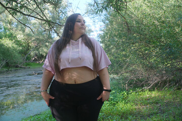 Plus size woman stands beside a quiet forest stream, wearing casual sportswear and gazing into the distance, expressing confidence, self acceptance and connection with nature © ullision