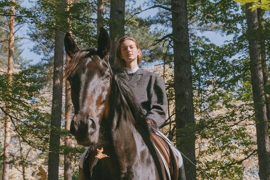 Young woman riding a dark brown horse through a sunlit forest trail with a calm expression for equestrian lifestyle and hobby content