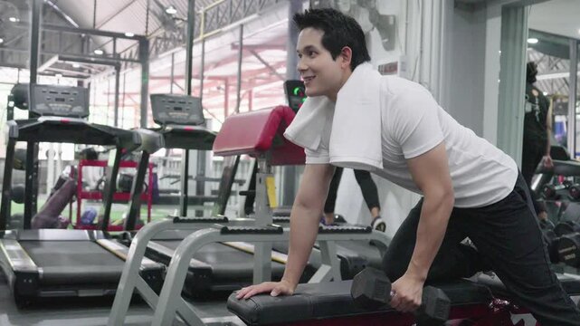 Handsome young Asian man doing single-arm dumbbell row exercise on a bench at the gym