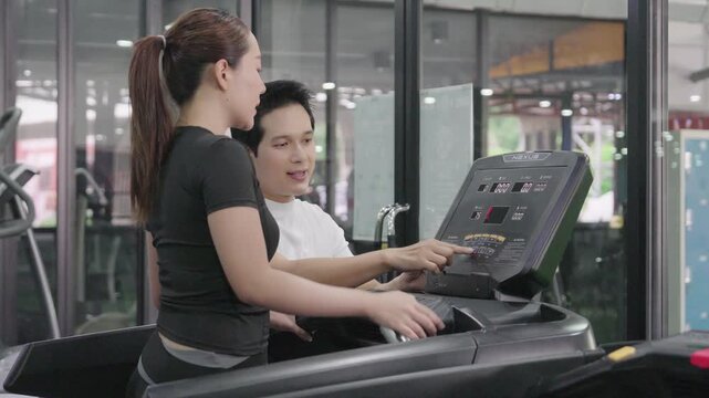 A friendly male trainer is setting up the treadmill display for an Asian woman exercising in a modern gym.