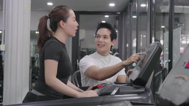 A friendly male trainer is setting up the treadmill display for an Asian woman exercising in a modern gym.