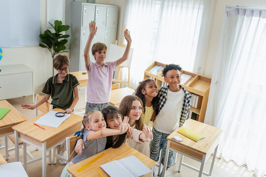 Diverse group of happy primary school children raising hands in classroom, Cheerful multi-ethnic students and teacher having fun in a modern classroom, Education concepts