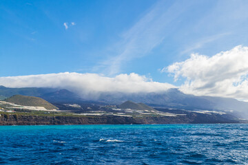 Obraz premium Volcanic cone of Tajogaite volcano visible on the horizon above clouds, shot from the sea near the lava delta in La Palma, Canary Islands, Spain