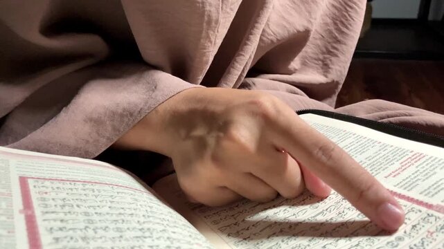 Vertical 4K slow motion pull out shot of a young muslim woman wearing hijab and glasses reciting holy Quran book with finger pointing verses Dramatic overhead lighting cozy spiritual indoor broll
