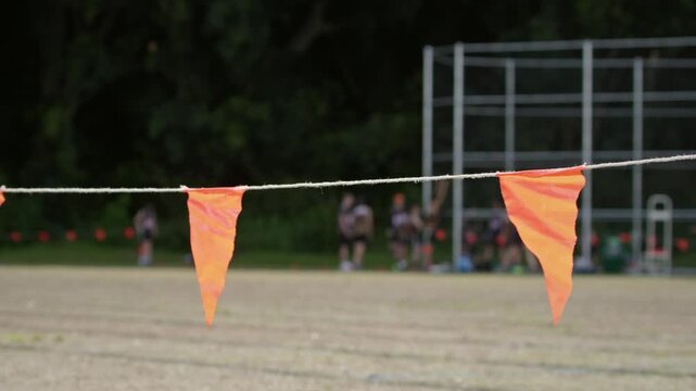 Orange boundary flags mark the edge of an athletics field during a junior sporting event, with athletes and spectators blurred in the background. Shallow depth of field. Medium shot.