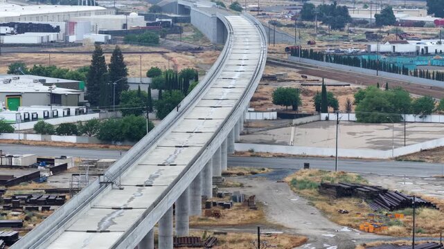 Cedar Viaduct high-speed rail deck stretches diagonally above a dry construction yard with steel materials and heavy equipment scattered below in Fresno, California, USA. Aerial drone view.