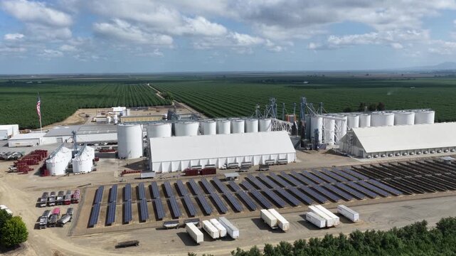 Almond processing facility with grain silos, solar panels and warehouse buildings surrounded by vast green orchard rows under dramatic clouds in Arbuckle, California, USA. Aerial drone rise wide