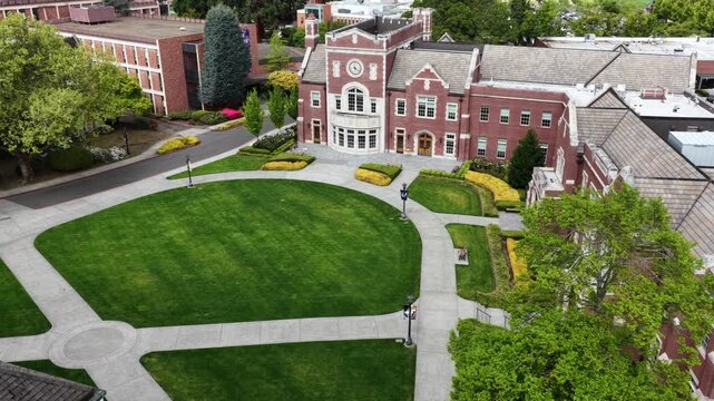 Historic red-brick university building with clock tower and manicured oval lawn anchors lush campus in Portland, with mature trees and academic buildings. Oregon, USA. Aerial drone view.