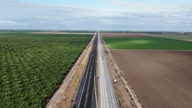 Empty highway cuts straight through patchwork farmland, lush green crop rows bordering freshly tilled soil beneath partly cloudy blue sky. Chowchilla, California, USA. Aerial drone rise wide shot
