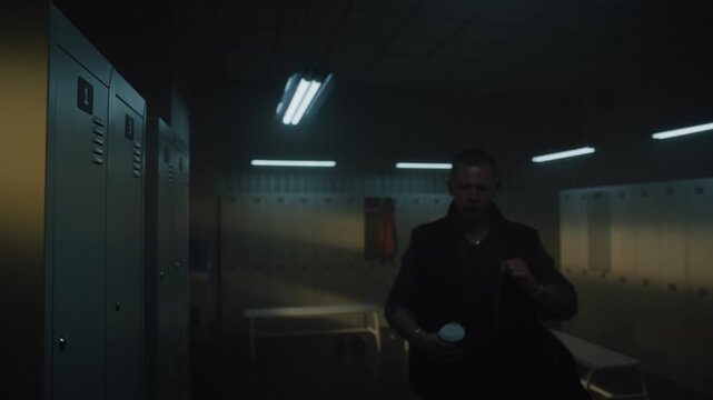 Man With Slight Limp Taking Off Vest, Enjoying Takeaway Coffee While Leaning on Locker and Looking Pensively Toward Window in Modern Sports Facility. Concept of Morning Routine, Authentic Lifestyle.