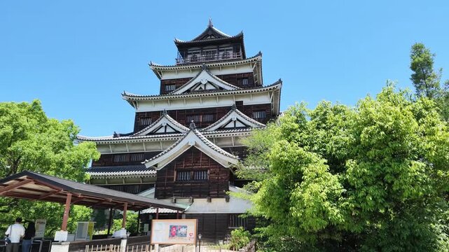 A scenic view of the historic Hiroshima Castle main tower surrounded by lush green trees under a clear blue sky in Japan