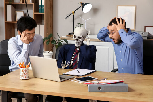 Stressed male colleagues with human skeleton at table in office