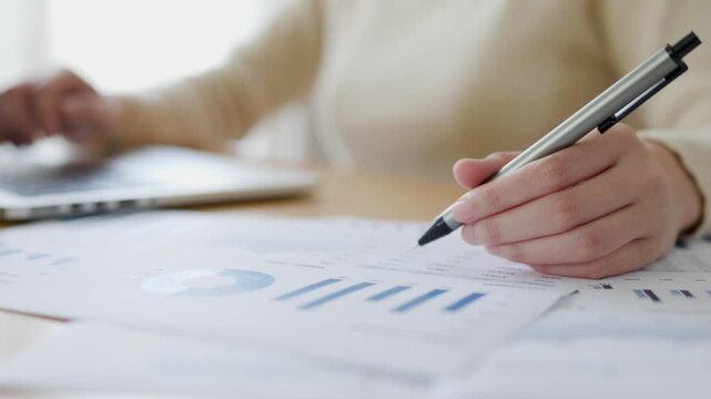 Close-up of a person analyzing printed financial charts with a pen, working at a desk beside a laptop