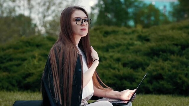 Young woman sits outdoors on grass, laptop open. She wears glasses and dark jacket, focused on screen. Green bushes surround her, natural daylight. Calm, studious mood. Ideal for remote work