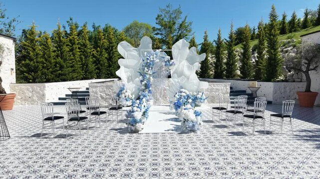 Elegant outdoor wedding setup with blue-white floral arch. Chairs arranged for guests around the central stage. Clear sky above lush green trees. Perfect for romantic ceremonies or upscale events
