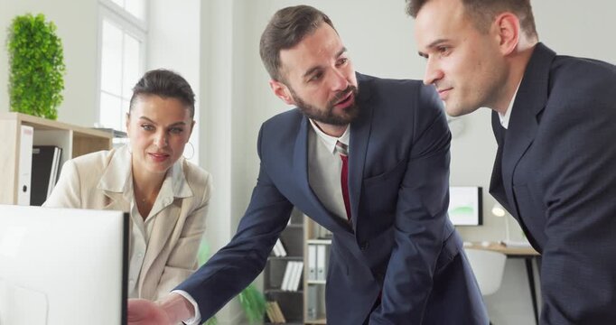 Managers team discussion around computer strategy. Three colleagues in formal suits review data at a monitor, aligning goals in a bright office. Strong concept of collaborative business planning.