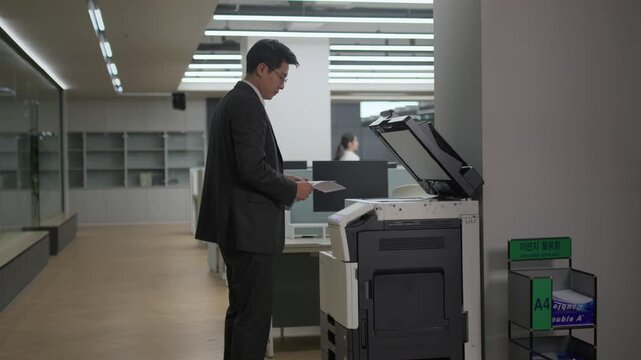 A businessman in a suit standing at a printer or copier machine in a bright modern office while working late at night.