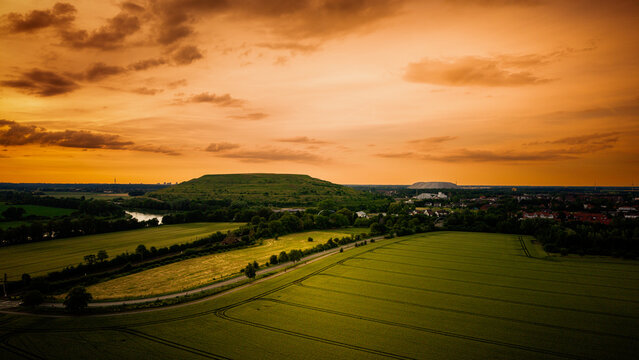Aerial view of green agricultural fields, a winding river, and a prominent industrial mound under a vibrant golden sunset sky in Sehnde, Lower Saxony, Germany.