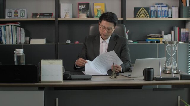 A businessman in a suit sitting at a modern executive desk reviewing documents with a laptop and bookshelf in the background late at night.