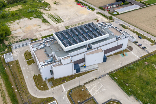 Aerial view of an advanced AI datacenter featuring a large cooling system on the roof, surrounded by paved parking areas and green fields in Warsaw, Masovian Voivodeship, Poland.