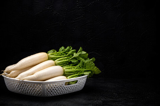 Fresh white daikon radishes with green leaves in a white basket on a black background.