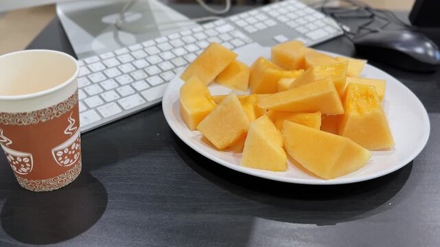 Close-up of fresh orange cantaloupe melon cubes on a white plate next to a paper coffee cup. The healthy office snack rests on a dark modern workspace desk beside a white computer keyboard.