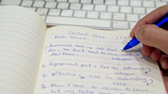 Close-up of a hand using a blue pen to write social media content ideas in a lined notebook. The content planner sits on a wooden desk next to a blurred white keyboard in a bright office workspace.