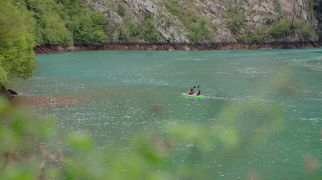 Two people kayaking across turquoise waters of Lake Dajes, Slovenia, surrounded by forested shores and rocky hillsides. Static view