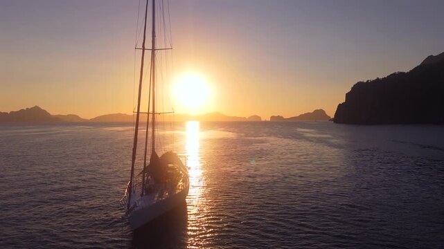 Close drone approach toward an anchored sailboat silhouetted against the golden El Nido sunset
